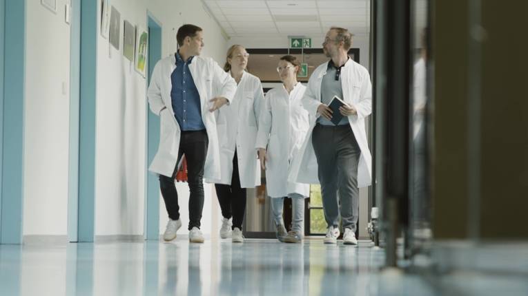 Two men and two women in white coats walk down a corridor.